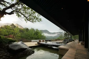 Modern Japanese-style garden with stone-lined pond, wooden deck, white outdoor sofas, and lush greenery overlooking misty mountains.
