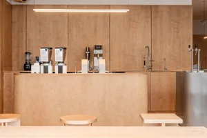 Modern minimalist coffee bar made of light birch plywood with built-in espresso machines, grinders, and stainless steel taps, illuminated by a long linear pendant light.