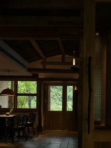Interior of a Japanese-style countryside house dining room featuring dark wood beams, wooden dining table with black rattan chairs, large windows overlooking a green forest, and warm pendant lighting.