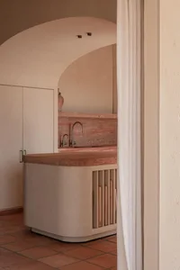 View through a linen curtain into a modern kitchen featuring a curved island with terracotta countertop, plaster base, and wooden slat storage, set against a large arched opening and terracotta floor tiles.