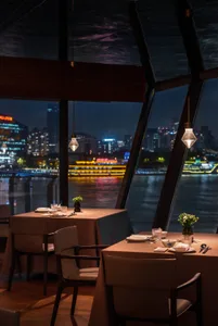 Elegant modern restaurant interior at night featuring two set dining tables with beige tablecloths and leather chairs, hanging glass pendant lights, dark wooden ceiling beams, and a stunning nighttime view of a river and illuminated city skyline through floor-to-ceiling angled windows.