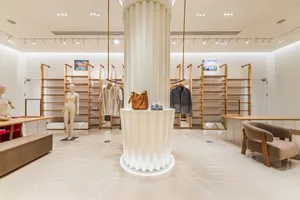 Modern minimalist clothing boutique interior with a large white fluted column display pedestal, wooden clothing racks, mannequins, and beige upholstered armchair on a polished marble floor.
