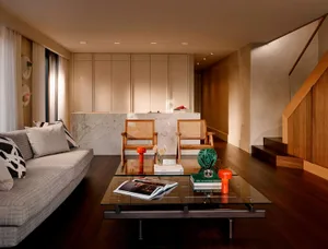 Modern living room featuring a light gray sofa, glass coffee table with books and orange lamps, two rattan armchairs, a white marble kitchen island in the background, and a wooden staircase on the right.