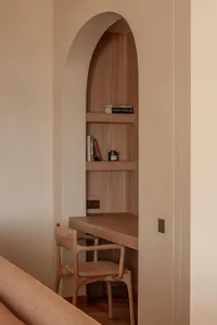 Built-in oak desk and chair placed within a tall arched niche in a neutral-toned wall, with books and a candle on the shelves.
