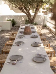 Long outdoor dining table made of light stone with woven rush stools, set with ceramic plates and a large terracotta pot centerpiece in a gravel courtyard under an olive tree.