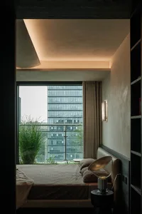 Modern minimalist bedroom featuring a concrete ceiling and walls, large floor-to-ceiling window overlooking a city skyline and green balcony, neutral-toned bedding, and a sculptural metallic table lamp.