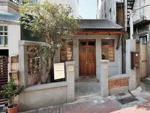 Street view of a renovated old house entrance featuring a grey concrete wall, wooden double door, small tree, and brick pavement in a narrow alley.