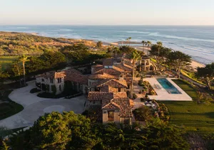 Aerial view of a luxurious Mediterranean-style stone villa with terracotta tile roofs, swimming pool, and manicured gardens overlooking the Pacific Ocean at sunset.