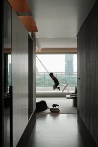 Modern minimalist apartment interior featuring a child on an indoor swing, concrete walls, large glass balcony overlooking a city skyline, and scattered children's toys on a light rug.