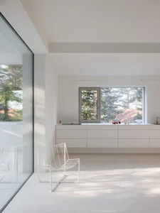 A bright, minimalist living room with white walls, a white wireframe chair, floating white cabinetry, and large windows overlooking green trees and a tiled roof.