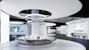 Modern futuristic exhibition hall with a large circular white platform displaying a detailed architectural city model, surrounded by concentric black and white lighting rings hanging from the ceiling, digital screens on curved walls, and minimalist white and dark grey interior.