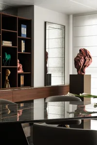 Modern dining room featuring a dark walnut built-in cabinet displaying Chinese art pieces, a large textured mirror, and a dramatic red stone sculpture on a pedestal, with a glossy black dining table in the foreground.