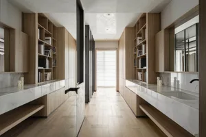 Modern minimalist walk-in closet with light oak built-in shelving, white marble countertops, and a central dressing aisle leading to a bright window.