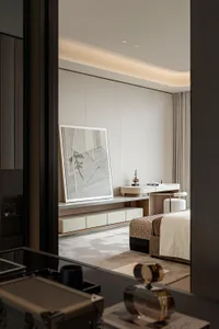 View into a minimalist luxury bedroom with neutral tones, a woven ottoman bench at the foot of the bed, a floating vanity desk with upholstered stool, and a large framed abstract line drawing on the wall.