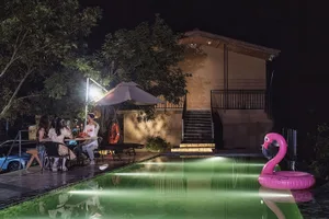 夜晚的現代泳池露台，三人圍坐在餐桌旁用餐，水下泳池燈光投射出綠色光暈，大型粉紅火鶴充氣浮圈，搭配LED照明的木質階梯與現代建築背景。