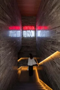 Woman standing on a modern stone staircase looking at mirrored red and white neon signs that read I CAN'T GO ON and I'LL GO ON
