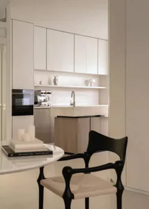 Modern minimalist kitchen with white matte cabinetry, light wood breakfast bar, built-in oven, open shelving, and a sculptural black chair in the foreground.