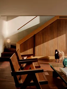 Two wooden armchairs with cane backs placed under a wooden staircase, next to a glass coffee table and a wool sheep sculpture in a sunlit modern interior.