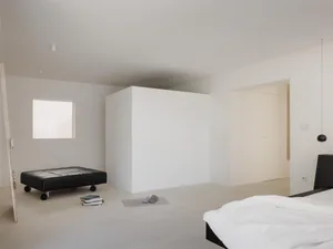Minimalist bedroom interior with white walls, polished concrete floor, black low platform bed with white bedding, black leather rolling ottoman, and a small square window providing natural light.