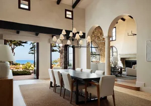 Spacious coastal Mediterranean dining room featuring a long dark wood table, eight upholstered chairs, exposed black ceiling beams, large arched openings revealing ocean view, and a stone fireplace in the background.