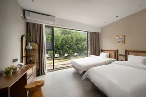 Modern minimalist twin hotel room featuring two beds with wooden and rattan headboards, a large floor-to-ceiling window overlooking a bamboo forest and outdoor tents, wooden desk with live-edge stool, and neutral beige walls.
