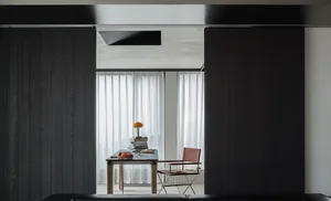 Modern minimalist home office viewed through dark wood sliding doors, featuring a blue desk with vintage red telephone, stacked books, an orange mushroom lamp, and a red leather director’s chair.