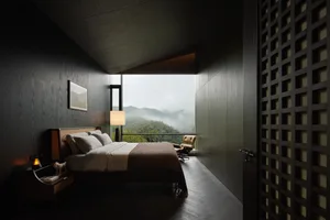 Modern dark wood bedroom interior featuring a large bed with neutral linens, a floor lamp, and a wide opening revealing a misty mountain and forest view.