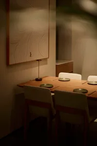 Modern minimalist dining room featuring a solid oak table, four upholstered chairs with wooden frames, a sculptural table lamp, and a large framed Japanese line-drawing wall art with calligraphy.