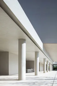 Modern brutalist outdoor colonnade featuring evenly spaced concrete columns under a massive concrete beam with strong sunlight casting long shadows.