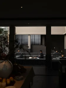 Night view of a modern minimalist concrete courtyard with built-in grey sofas, a man sitting in the sunken lounge area, warm candlelight, and dark architectural pillars.