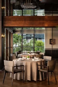 Elegant modern restaurant interior featuring round dining tables with beige tablecloths, walnut wood beams, leather-upholstered chairs, and large windows overlooking green trees and city buildings.
