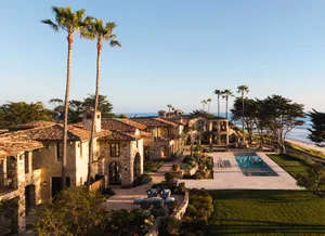 Aerial view of a luxurious stone villa with terracotta tile roofs, multiple chimneys, lush landscaping, a rectangular swimming pool, and direct oceanfront access with palm trees and the Pacific Ocean in the background.
