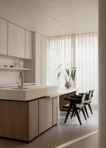 Modern minimalist kitchen featuring a large oak island with integrated sink, white upper cabinets, and three black dining chairs with cane backs placed beside a long table near a window with sheer curtains.
