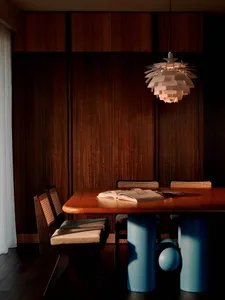 Modern dining room featuring a walnut dining table with bright blue cylindrical pedestal legs, cane-back dining chairs, and a large white Artichoke pendant lamp hanging above an open book on the table.