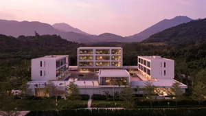 Aerial view of a contemporary concrete resort complex with illuminated interiors, surrounded by tropical landscaping and dramatic mountains at twilight.