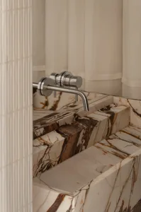 Close-up of a luxurious Calacatta marble vanity sink with dramatic brown veining and a modern brushed nickel wall-mounted faucet against a neutral textured wall.