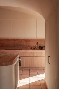 Modern minimalist kitchen featuring terracotta tiles, travertine countertops, light wood cabinetry, and a curved plaster archway with warm natural light.