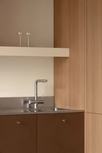 Close-up of a modern kitchen sink area with stainless steel faucet, integrated stainless steel backsplash and sink, warm wood lower cabinets with round pulls, and a floating white shelf above with two glass tumblers.