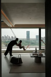 Modern minimalist living room featuring a suspended indoor swing, concrete ceiling, dark wood coffee table, and large floor-to-ceiling windows overlooking a city skyline with a father and son playing.