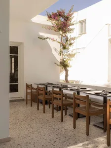 Outdoor courtyard dining area with wooden chairs, black marble tables, terrazzo floor, and a blooming bougainvillea tree under a bright blue sky.