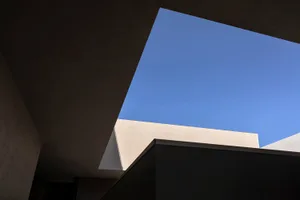 Low-angle architectural view looking up through a concrete building structure toward a clear blue sky, showing minimalist geometric forms and material textures.