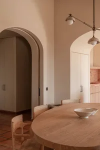 Minimalist dining area featuring a round solid wood table, light oak chairs, terracotta floor tiles, and soft beige arched doorways with warm pendant lighting.