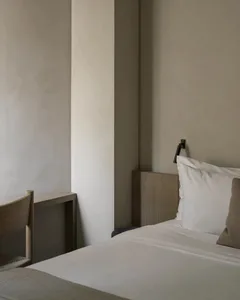 Minimalist bedroom corner featuring a light oak headboard, white bedding, textured beige pillow, black wall sconce, and a partial view of an oak chair against a textured neutral wall.