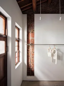 Interior corner of an industrial loft featuring an exposed red brick wall, white painted surfaces, wooden ceiling beams, a metal clothing rail with white garments, and natural light from a wooden window.