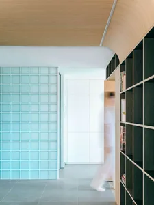 Modern minimalist hallway featuring a curved light-wood ceiling, translucent turquoise glass block wall, dark green built-in bookshelf, and grey tiled floor with a person walking by.
