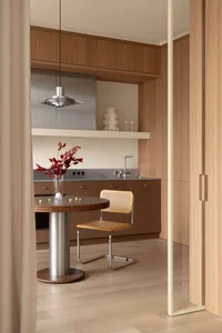 View of a modern minimalist kitchen dining area with walnut cabinetry, a round wooden dining table on a metal pedestal, a cane and leather Cesca-style chair, stainless steel countertop and sink, and a large metallic pendant lamp.