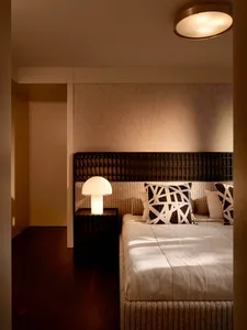 Modern minimalist bedroom with dark wood grid headboard, textured beige wallpaper with geometric line pattern, white mushroom table lamp, and black and white abstract pillows on a woven bed base.