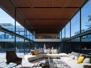 Modern double-height living room featuring a large L-shaped sectional sofa, brown leather armchair, glass coffee tables, and dramatic wood slat ceiling with full-height glass walls overlooking a courtyard.