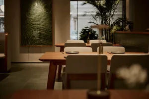 Modern minimalist restaurant dining area featuring light oak wooden tables, beige upholstered chairs, large textured green artwork on the wall, and abundant indoor plants near large windows.