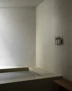 Minimalist bathroom corner featuring a sunken travertine bathtub with integrated stone ledge and wall-mounted soap dispensers.